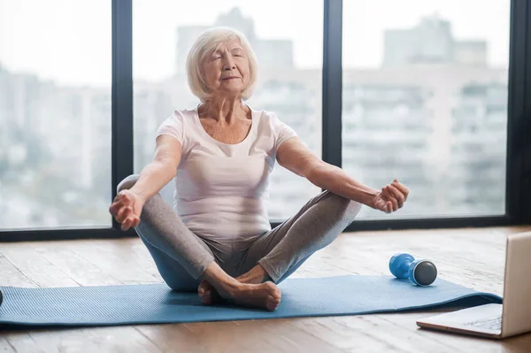 Grauhaarige Seniorin sitzt auf dem Boden und nimmt an einem Online-Yoga-Kurs teil — Stockfoto