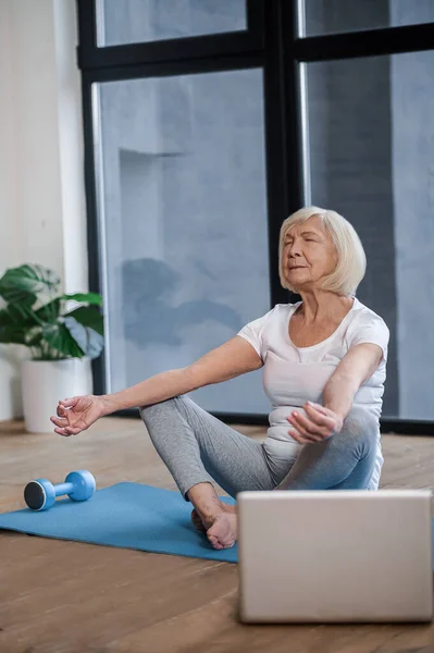 Gray-haired senior woman sitting on the floor and having an online yoga class — 스톡 사진