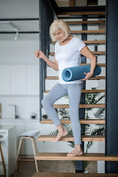 Eine Seniorin steigt mit einer Yogamatte in der Hand die Treppe hinunter — Stockfoto