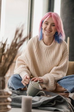 Pembe saçlı genç kız, pencerenin yanında çay içiyor.