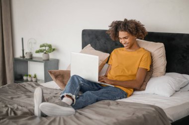 Yatakta dizüstü bilgisayarla oturan kıvırcık saçlı genç adam.