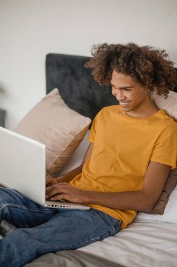 Yatakta dizüstü bilgisayarla oturan kıvırcık saçlı genç adam.