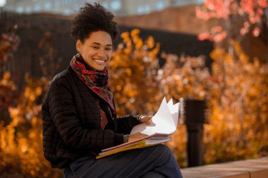 Elinde kitaplarla bankta oturan esmer tenli kız.