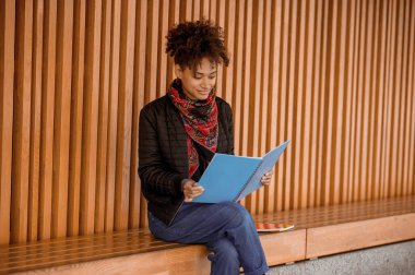 Elinde kitapla bankta oturan siyah ceketli bir kız.