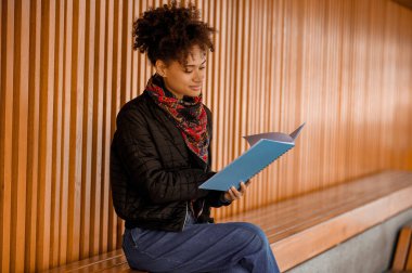 Elinde kitapla bankta oturan siyah ceketli bir kız.