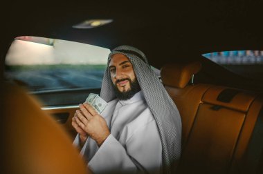 Suudi adam geleneksel kıyafetlerle arabada para tutuyor.