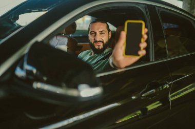 Genç, yakışıklı, siyah saçlı adam akıllı telefonunu gösteriyor.