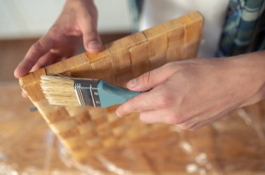 Marangoz ellerinin saman kutusunun kenarına boya fırçasıyla dokunduğu kesilmiş bir fotoğraf.
