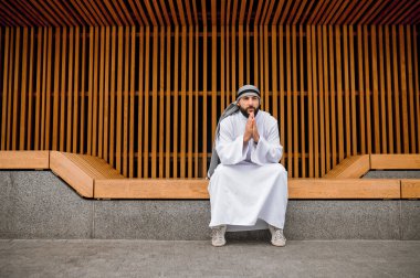 Dinleniyor. Ortadoğulu genç adam geleneksel giysiler içinde tahta bir bankta oturuyor.