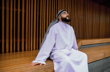 Dinleniyor. Ortadoğulu genç adam geleneksel giysiler içinde tahta bir bankta oturuyor.