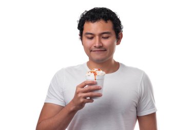 Genç bir İspanyol, Meksikalı, beyaz bir gömlek giyip beyaz bir arka planda gülümseyen, beyaz peynirli ve acı soslu bir bardakta mısır yiyor.