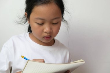 Beyaz tişörtlü Asyalı küçük kız. Evde, COVID-19 durumunda evden veya her yerden öğrenmek için kalem ve defter kullanıyor..