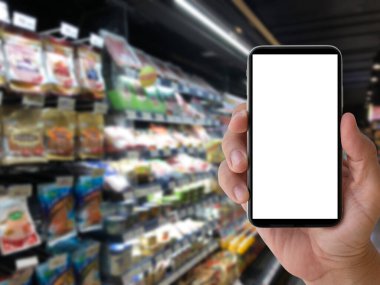 Bulanık süpermarket ya da bakkal arka planında beyaz ekranlı akıllı telefonu mesaj ya da sembol olarak kullanmak. İnternetten sipariş alışverişi