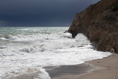 Fırtınalı kıyı şeridi, su sıçraması ve karanlık gökyüzü, turkuaz denizi olan kumlu sahil, Sitges, İspanya