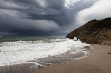 Fırtınalı kıyı şeridi, su sıçraması ve karanlık gökyüzü, turkuaz denizi olan kumlu sahil, Sitges, İspanya