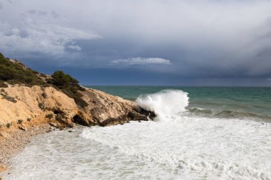 Fırtınalı kıyı şeridi, su sıçraması ve karanlık gökyüzü, turkuaz denizi olan kumlu sahil, Sitges, İspanya