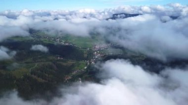 Dağlardaki bir köyün üzerindeki puslu bulutlar. Bulutların üstündeki kırsal dağ manzarasının görüntüsü. Hızlı hareket, yüksek hız, insansız hava aracı görüntüsü. 4K