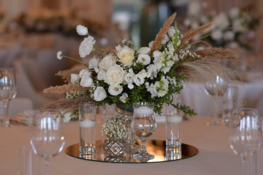 Beyaz masa örtüleri, temiz vazolar, beyaz kasımpatı ve eğrelti otu düzenlemeleri. Altın renkli tabaklar, peavh peçeteleri, masa numaraları ve ayna süslemeleri. Düğün günü.