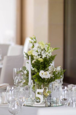 Beyaz masa örtüleri, temiz vazolar, beyaz kasımpatı ve eğrelti otu düzenlemeleri. Altın renkli tabaklar, peavh peçeteleri, masa numaraları ve ayna süslemeleri. Düğün günü.