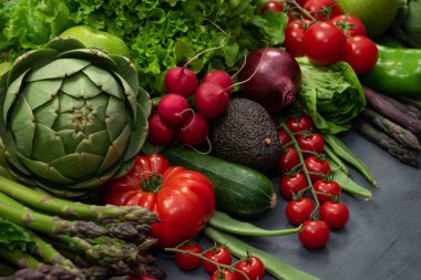 Farklı organik sebzeler - kuşkonmaz, domates vişne, avokado, enginar, biber, limon, limon, gri arka planda tuz. Fotokopi alanı olan yemek şöhreti, üst manzara. Çiğ vejetaryen konsepti