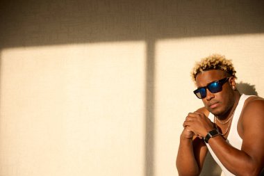 Güneş gözlüklü ve beyaz tişörtlü acımasız siyah adam güneş ışığının altında çömeliyor.