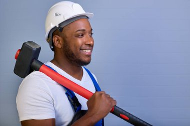 Beyaz kasklı ve iş elbiseli, dişlek bir gülümsemeyle gülümseyen, omzunda balyoz tutan memnun bir zenci. Tulum giymiş mutlu bir Afro-Amerikan işçisinin portresi.