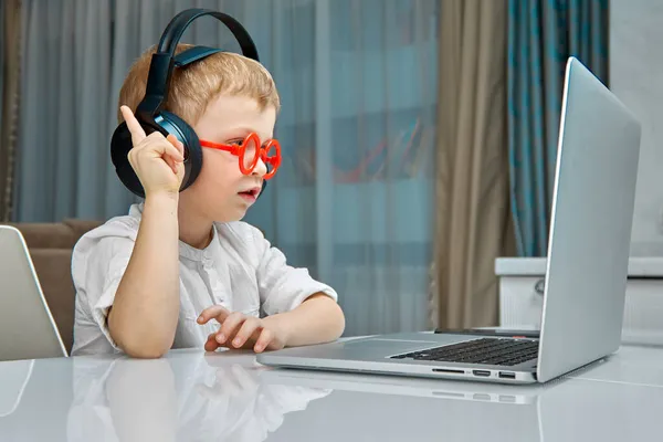 Ένα αγόρι με κόκκινα γυαλιά και μαύρα ακουστικά μελετά τη συσκευή laptop — Φωτογραφία Αρχείου