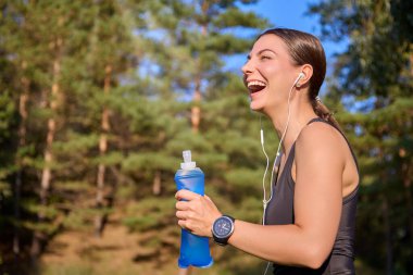 Fitness sporcusu. Genç bir kadın, güneşli bir günde parkta antrenman yaptıktan sonra kulaklıkla müzik dinliyor, neşeyle gülüyor ve tekrar kullanılabilir yumuşak bir su şişesinden su içiyor.