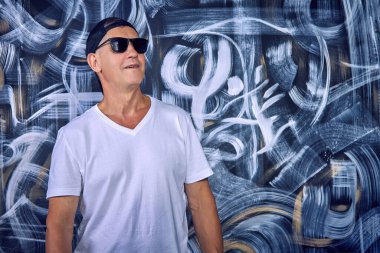 Beyaz tişörtlü, güneş gözlüklü ve duvarın karşısında duran havalı yaşlı bir adamın yakın çekimi. Emeklilik yaşında modern sporcu aktif kişi