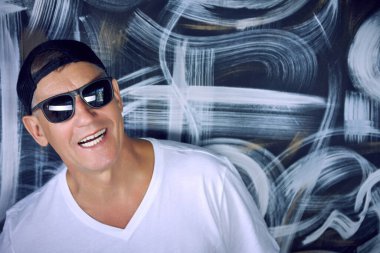 Beyaz tişörtlü, güneş gözlüklü ve duvarın karşısında duran havalı yaşlı bir adamın yakın çekimi. Emeklilik yaşında modern sporcu aktif kişi