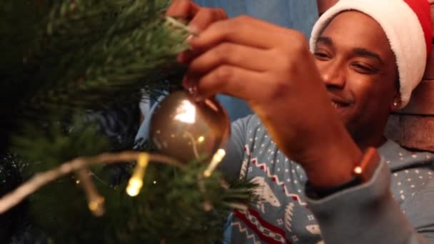 Fekete férfi pulóverben és kalapban karácsonyfát díszít. — Stock videók