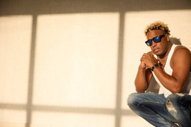 Güneş gözlüklü ve beyaz tişörtlü acımasız siyah adam güneş ışığının altında çömeliyor.