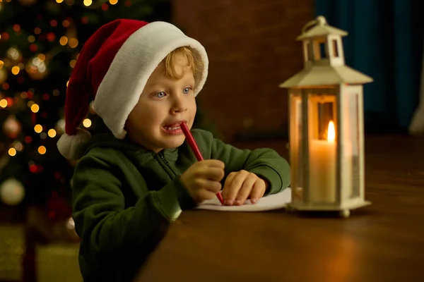 Boy in a Christmas hat writes a letter to Santa Claus by the light of a lantern at the Xmas tree