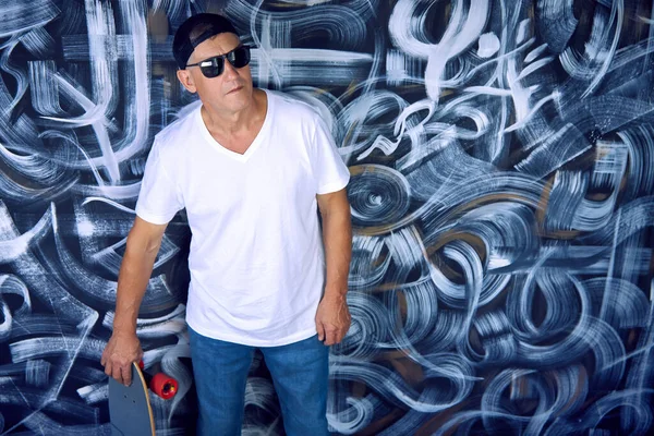 Hombre mayor fresco en una camiseta blanca, gafas de sol, una gorra y con un monopatín en las manos — Foto de Stock