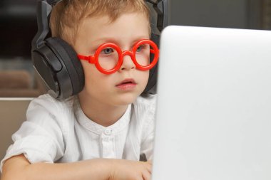 Kırmızı gözlüklü ve siyah kulaklıklı bir çocuk dizüstü bilgisayarı inceliyor.
