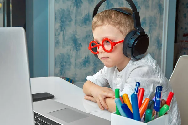 Ένα αγόρι που φοράει κόκκινα γυαλιά και ακουστικά παρακολουθεί περιεχόμενο σε ένα φορητό υπολογιστή — Φωτογραφία Αρχείου