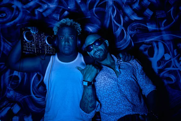 Un DJ afroamericano con una consola de DJ y su amigo latinoamericano en un club — Foto de Stock