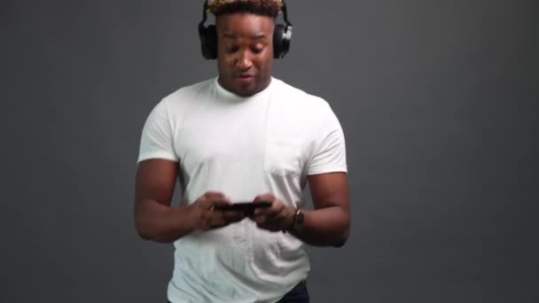 Enthusiastic African man with headphones is playing a game on a smartphone — Vídeo de Stock