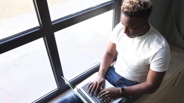 African-American man is browsing the Internet on laptop while sitting on window — Vídeo de Stock