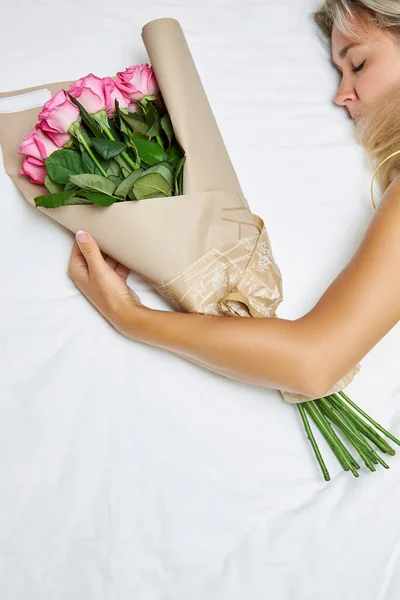 Dormir mujer se acuesta en la cama y abraza un ramo de rosas rosadas. — Foto de Stock