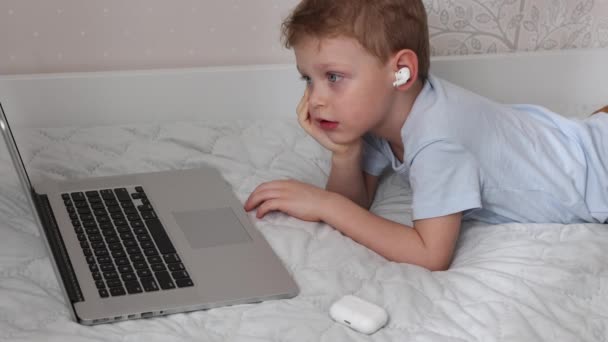 Kleiner blonder Junge mit drahtlosen Kopfhörern benutzt einen modernen Laptop auf dem Bett — Stockvideo