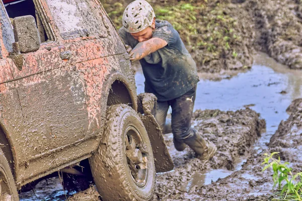 Homem usando um capacete empurra um carro off-road laranja 4x4 através da lama. — Fotografia de Stock