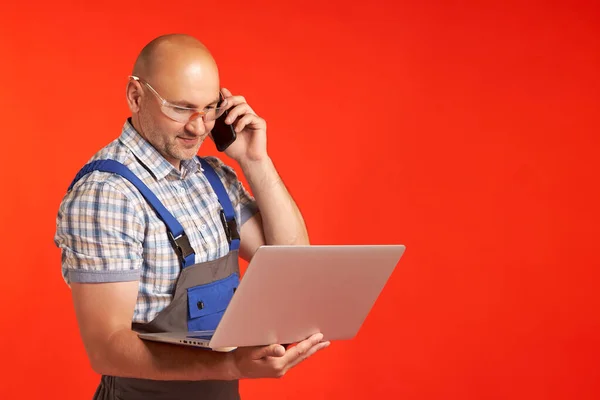 Skallig man i arbetskläder pratar i telefon och håller en bärbar dator — Stockfoto