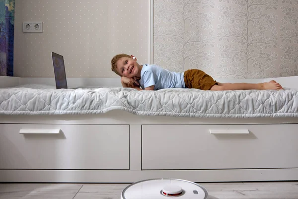 Jongen maakt gebruik van een laptop op het bed, terwijl de robot stofzuiger doet de reiniging — Stockfoto