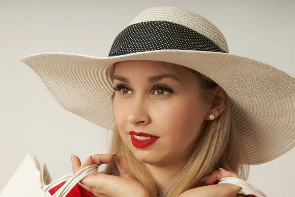 Hasır şapkalı, beyaz arka planda alışveriş torbalarıyla poz veren bir kadının yakın çekimi.