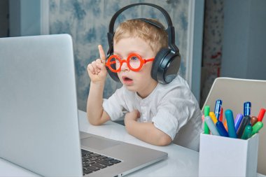 Kırmızı gözlüklü ve kulaklıklı bir çocuk bilgisayar kullanarak video bağlantısıyla iletişim kurarken eğleniyor..