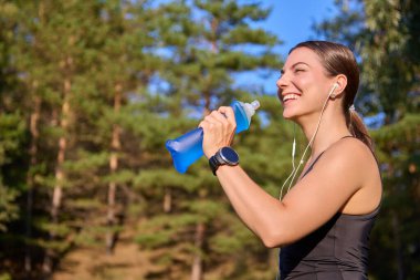 Genç bir kadın kulaklıkla müzik dinlerken gülüyor ve elinde yumuşak bir spor şişesi tutuyor.