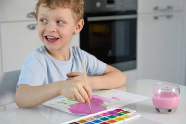 Sarışın, sevimli çocuk beyaz bir masada fırça ve pembe boya kullanarak resim yapıyor..