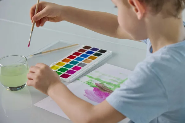 Pequeño niño sumerge un pincel en el agua y pinta en un pedazo de papel —  Fotos de Stock