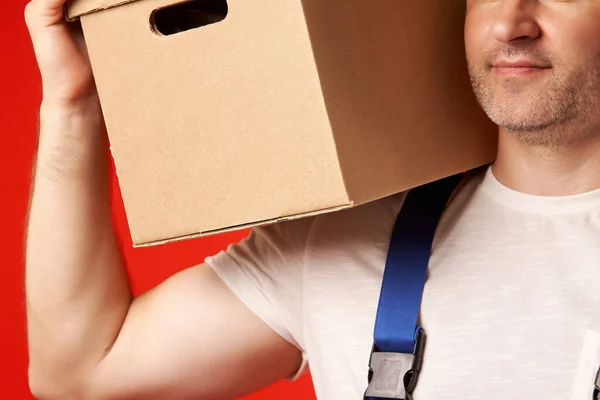 Bearded man in a loaders work suit holds a cardboard box on his shoulder, on a red background. A strong loader holds the box with his hand and shows his biceps. Physical training of the loader